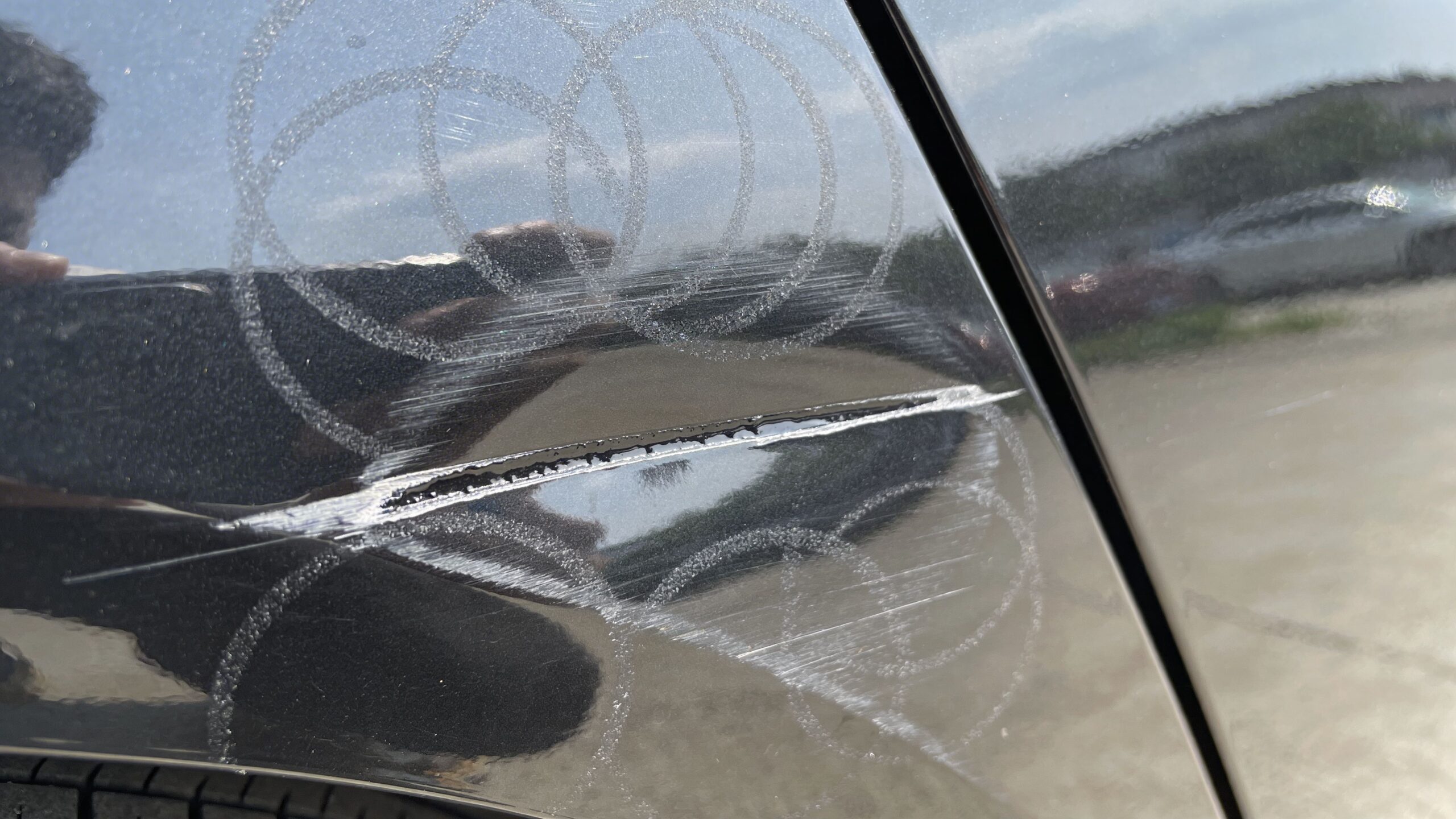 A deep, long scratch with surrounding circular swirl marks on the black paint of a car door.
