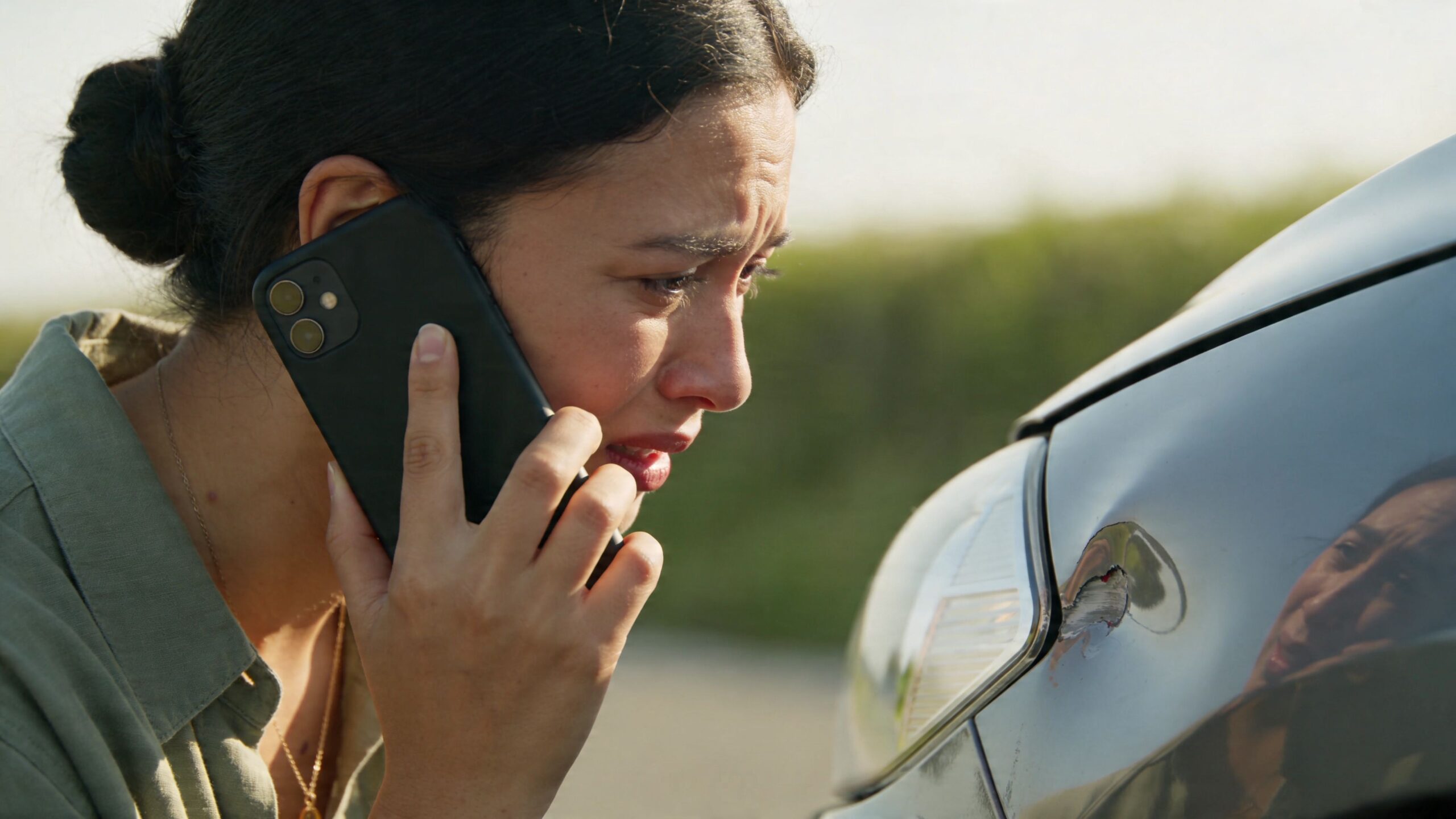 A distressed young woman making a phone call next to her car with a front bumper dent.